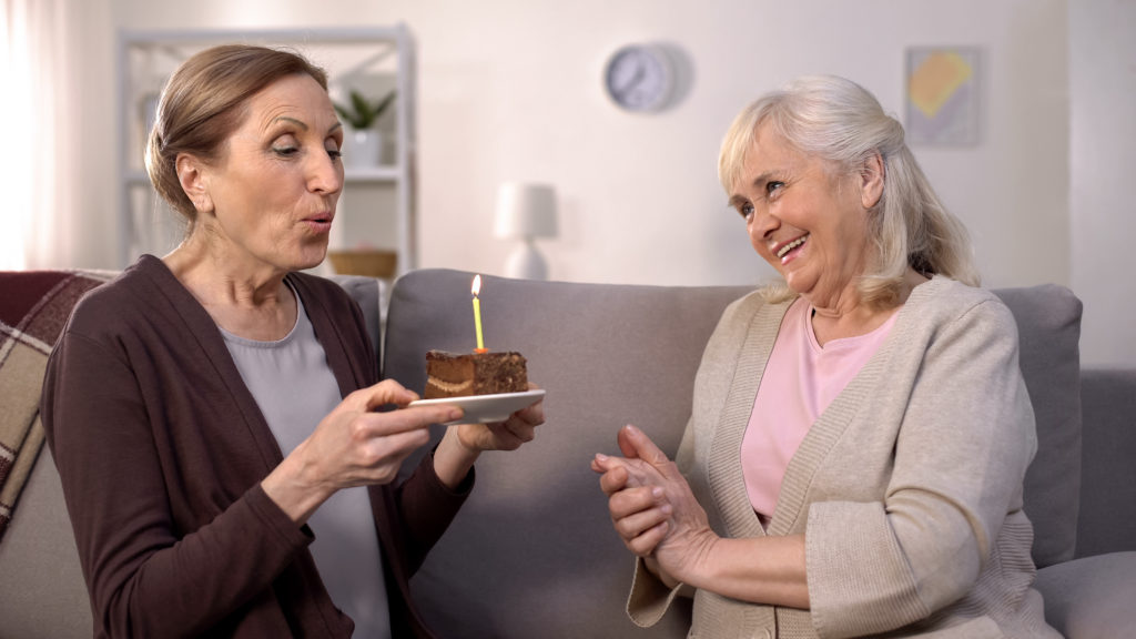 Mature woman blowing birthday cake candle sitting with smiling aged friend sofa